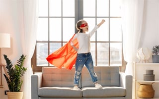 A small child dressed as a superhero standing, heroic, on a couch