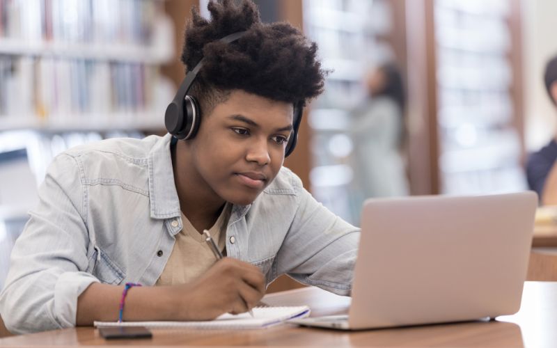 A young student with headphones studying with a laptop at a library