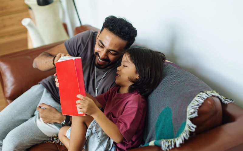 A parent and child looking at a book smiling on a couch.