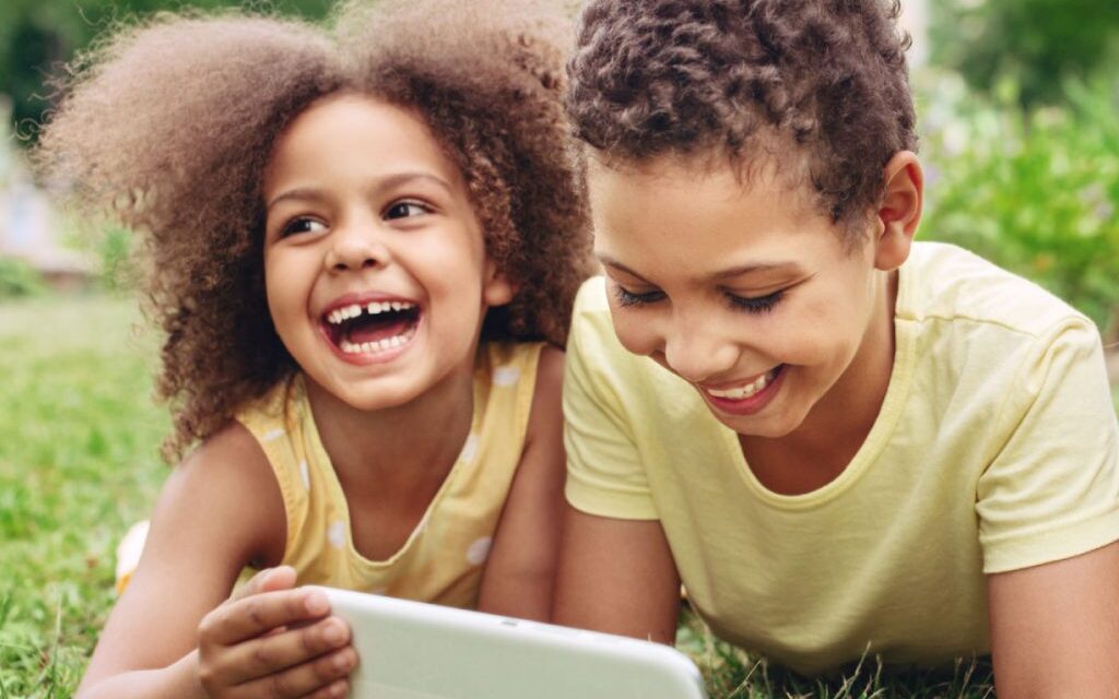 two kids with a tablet laughing on a lawn